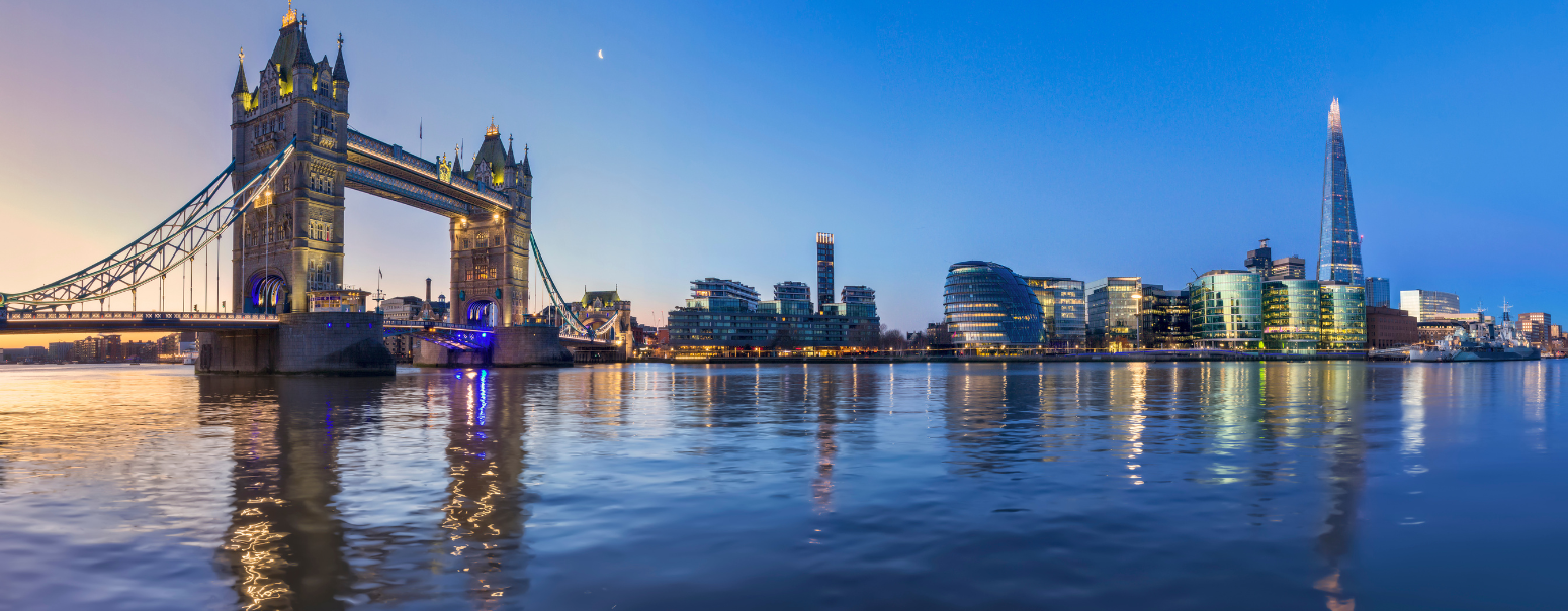 An image of Tower Bridget at dusk. The sky is blue and the water is a dark blue colour with the lights refracting on it.